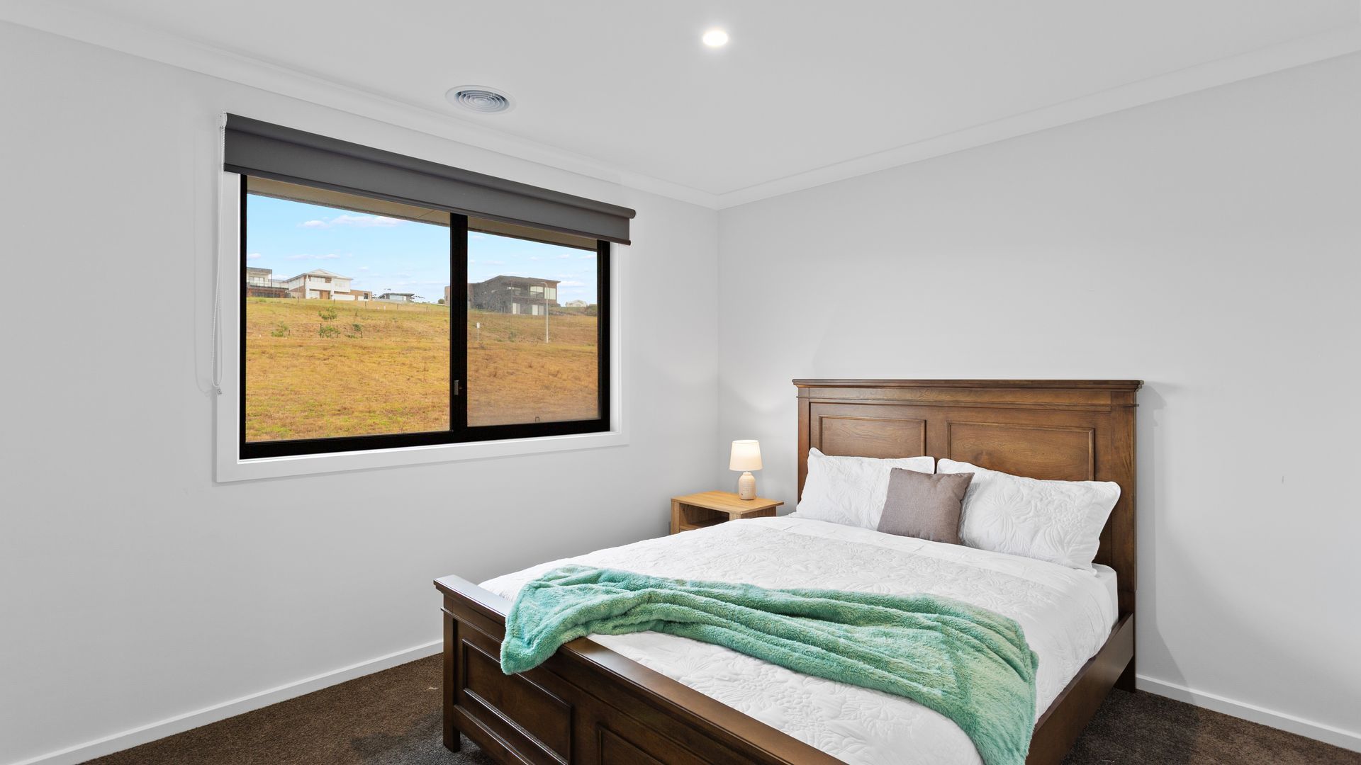 A bedroom with a large window, a wooden bed, a bedside table with a lamp, and a view of a field and buildings.