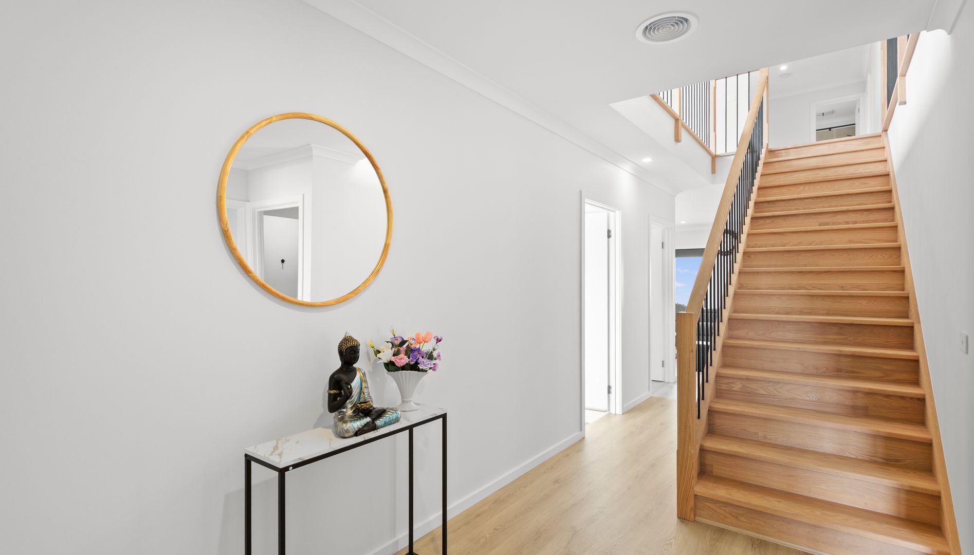 Ein modernes Interieur mit einer Holztreppe, einem runden Spiegel und einem kleinen Konsolentisch mit Buddha-Statue und Blumen.