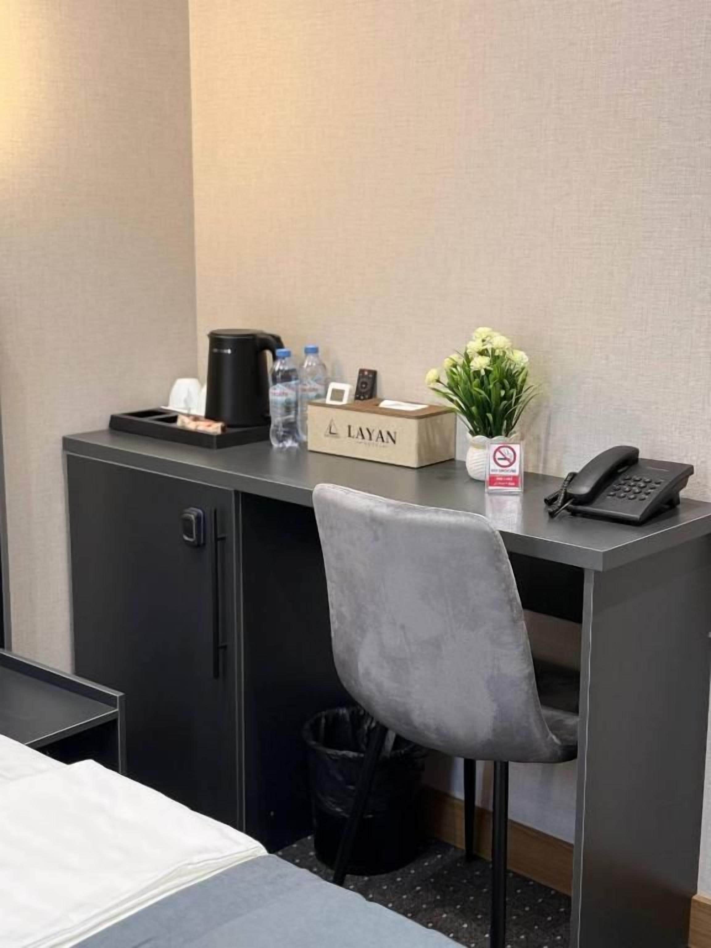 Habitación de hotel con escritorio, silla, cama, cubo de basura, botella de agua, cafetera, una caja etiquetada 'LAYAN', un florero con flores y un teléfono.