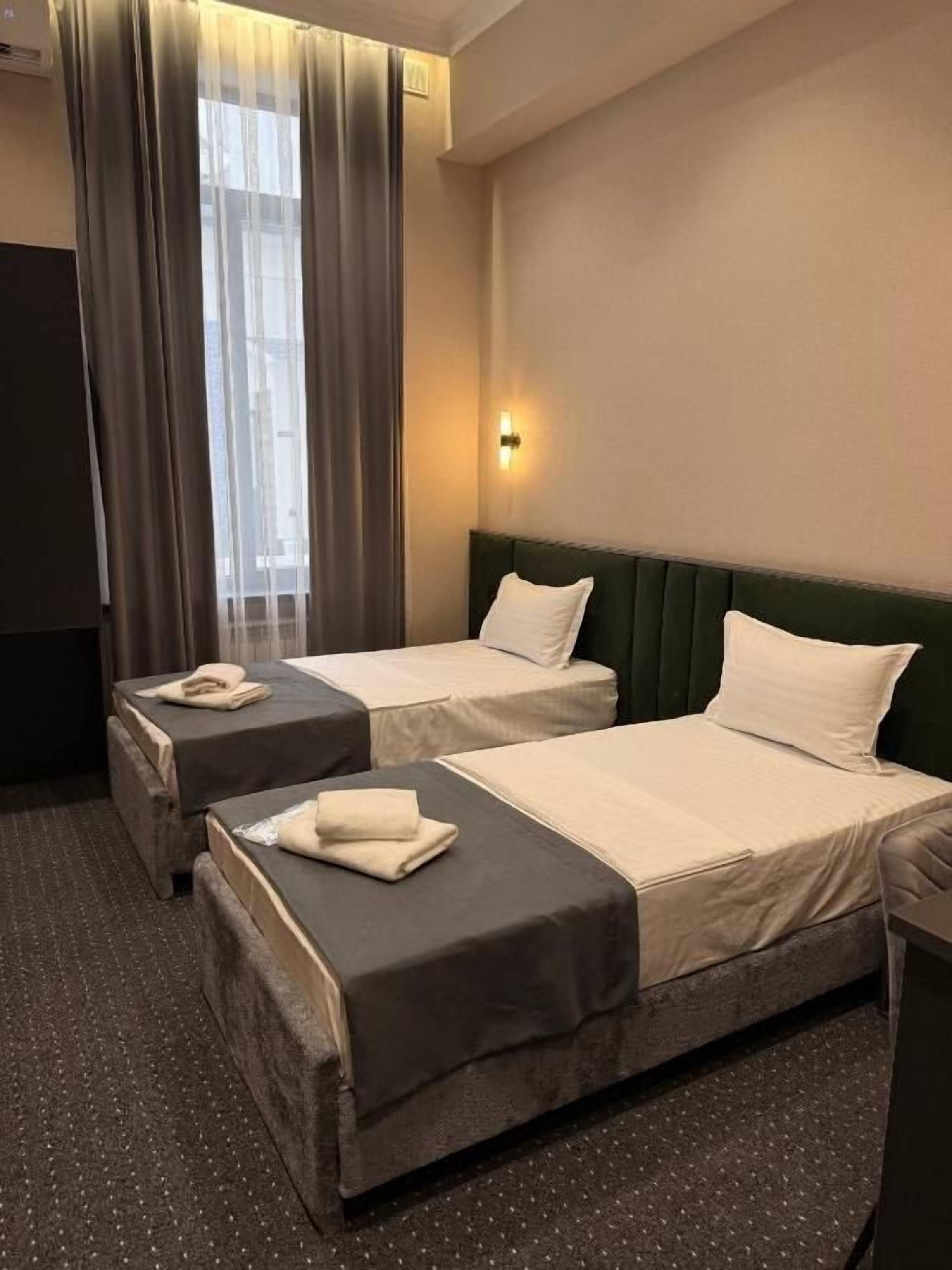 A hotel room with two beds, a green headboard, a wall-mounted light, and a window with curtains.