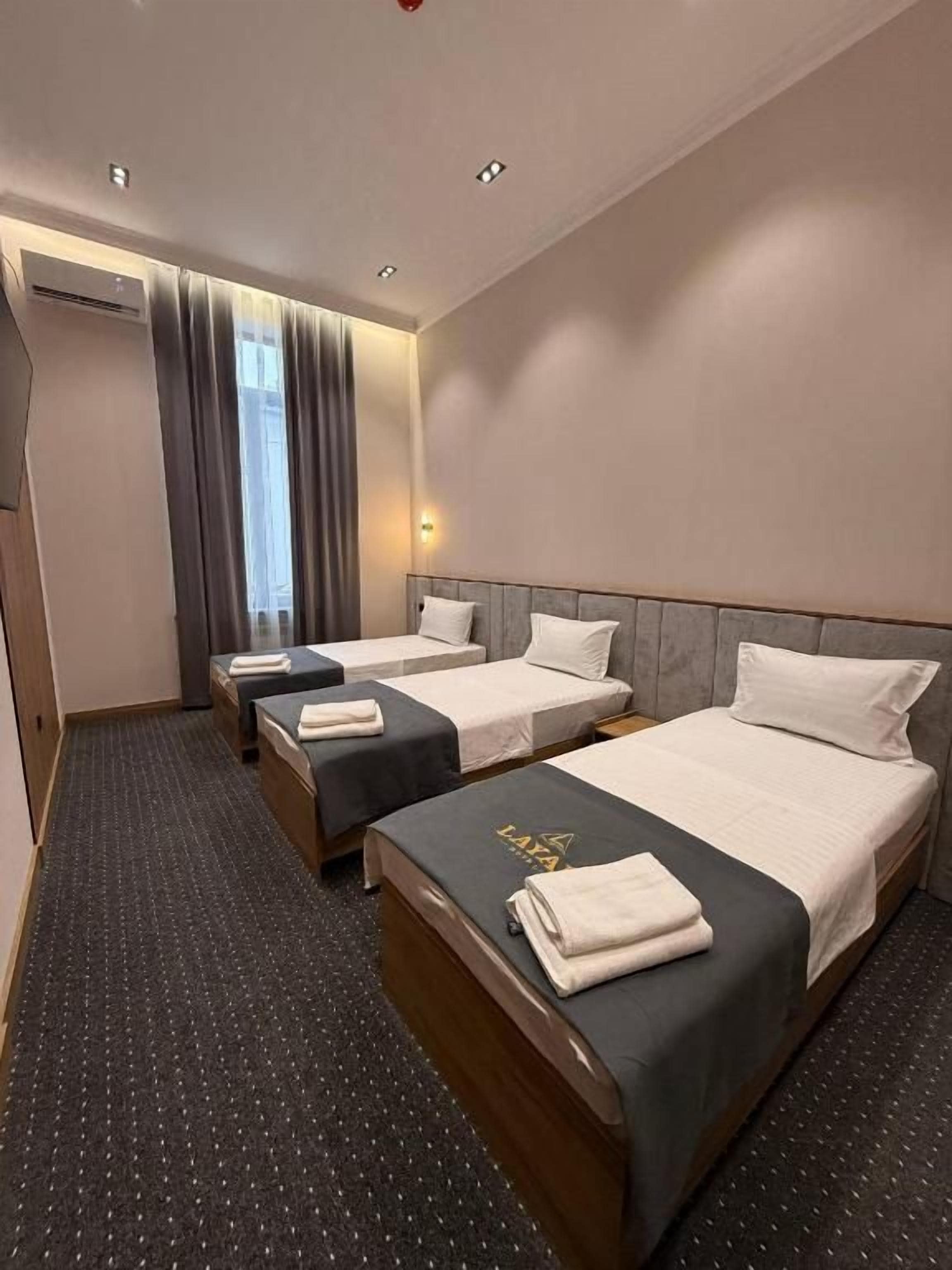 A hotel room with three beds, a large window with curtains, a carpeted floor, and a wall-mounted air conditioning unit.