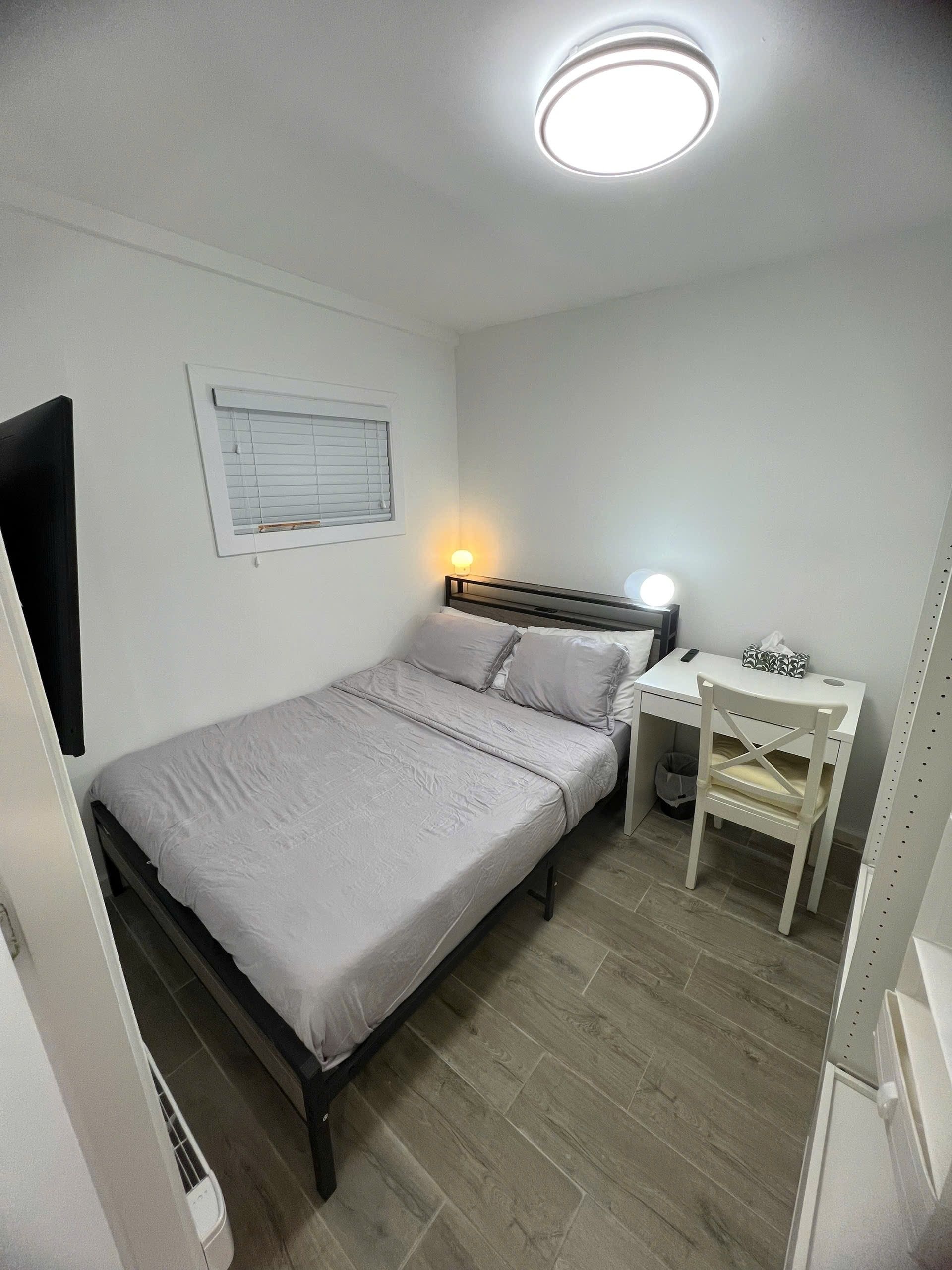 A bedroom with a bed, a desk, a chair, a window with blinds, and a ceiling light.