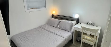 A bedroom with a bed, a desk, a chair, a window with blinds, and a ceiling light.