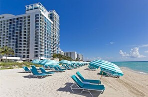 A beachfront hotel with multiple balconies, a sandy beach, and rows of blue and white striped beach umbrellas.