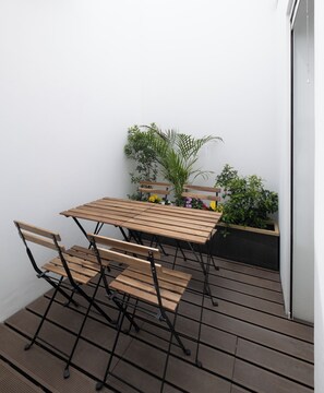 Une petite terrasse extérieure avec une table et des chaises en bois, entourée de plantes en pot et d’un mur blanc.