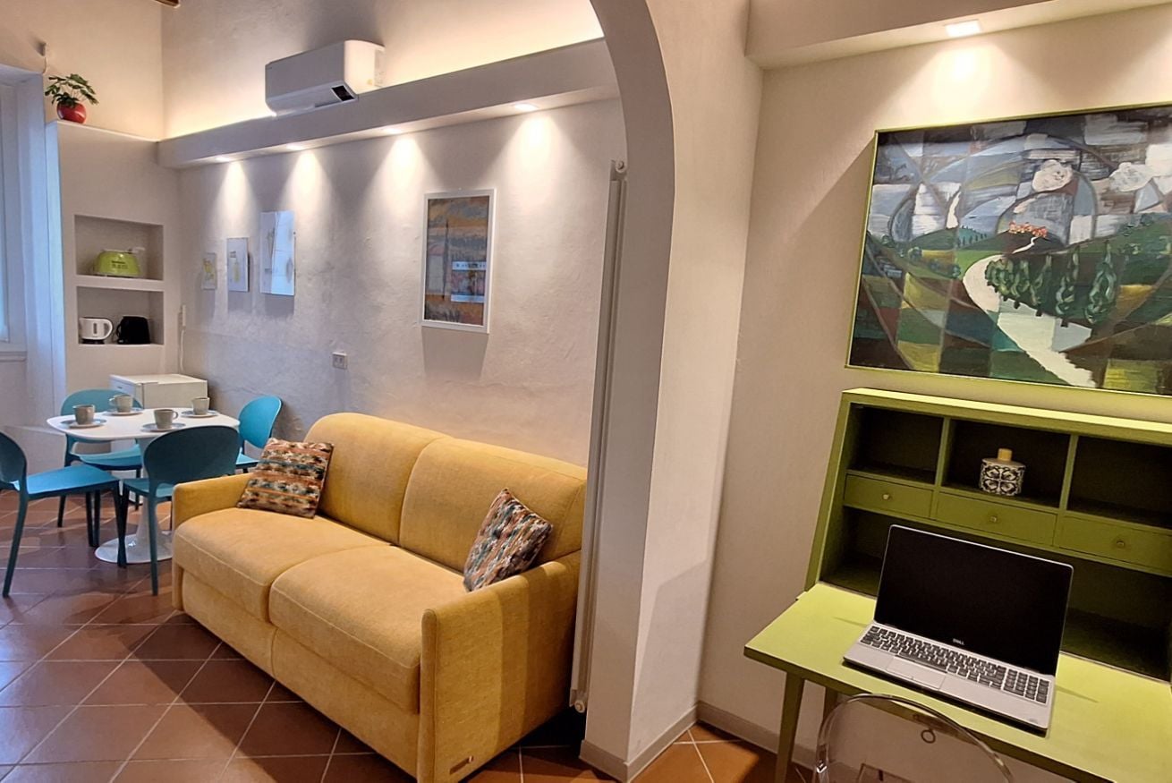 Ein Wohnzimmer mit gelber Couch, einem grünen Schreibtisch mit Laptop, einem Bild an der Wand und einem Essbereich mit Stühlen und Tisch.