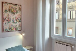 Una habitación con una cama, una mesita de noche, un radiador y una ventana con vistas a edificios.