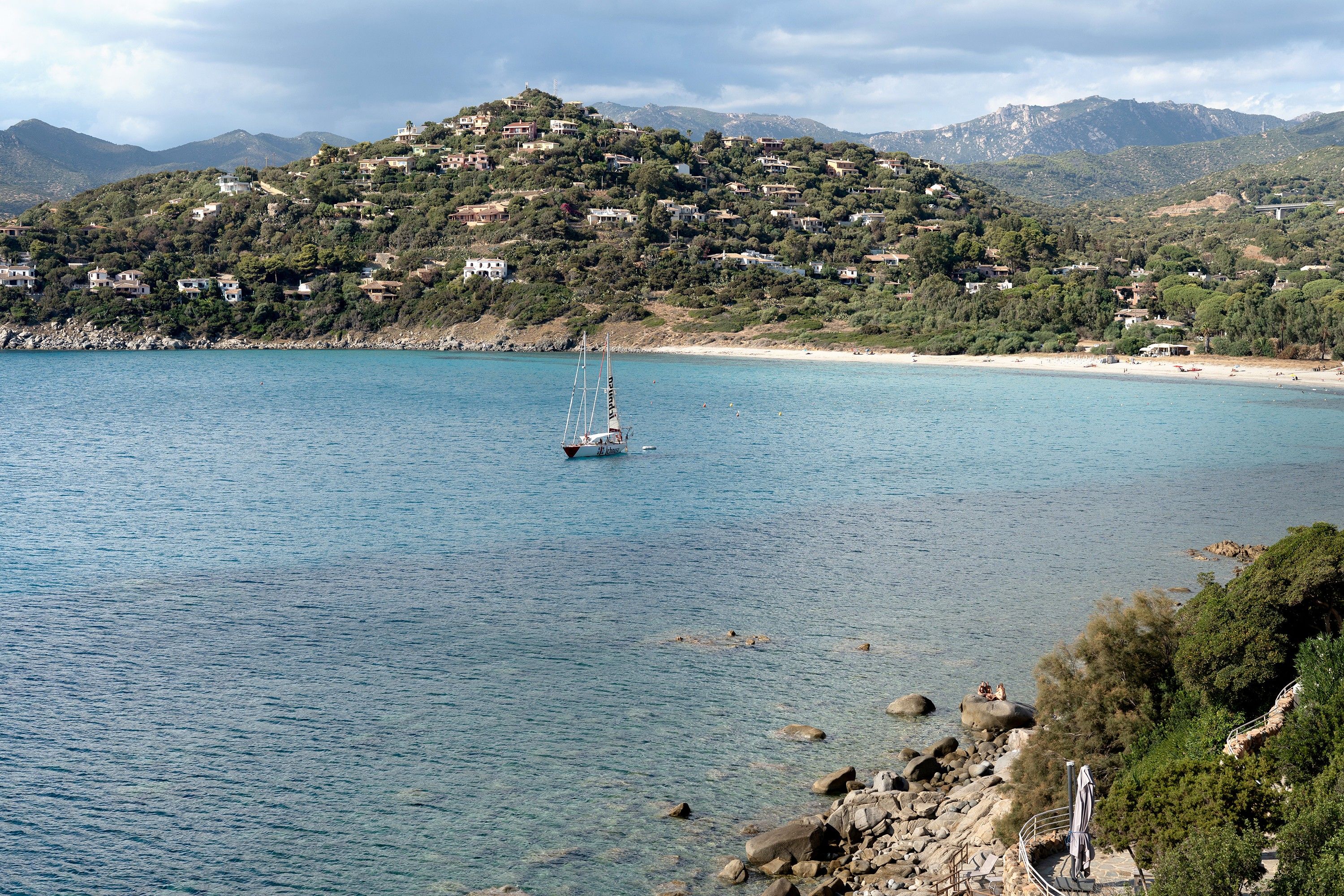 A coastal town with a clear view of the sea, a sailboat, and a hillside with buildings.