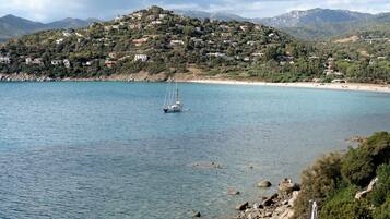 A coastal town with a clear view of the sea, a sailboat, and a hillside with buildings.