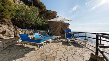 Une terrasse en bord de mer avec un sol en pierre, des fauteuils bleus et un parasol blanc.