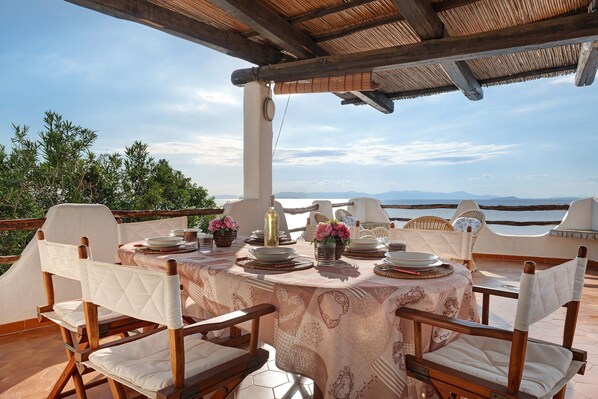 An outdoor dining area with a table set for four, surrounded by wicker chairs, under a wooden pergola with a view of the landscape.