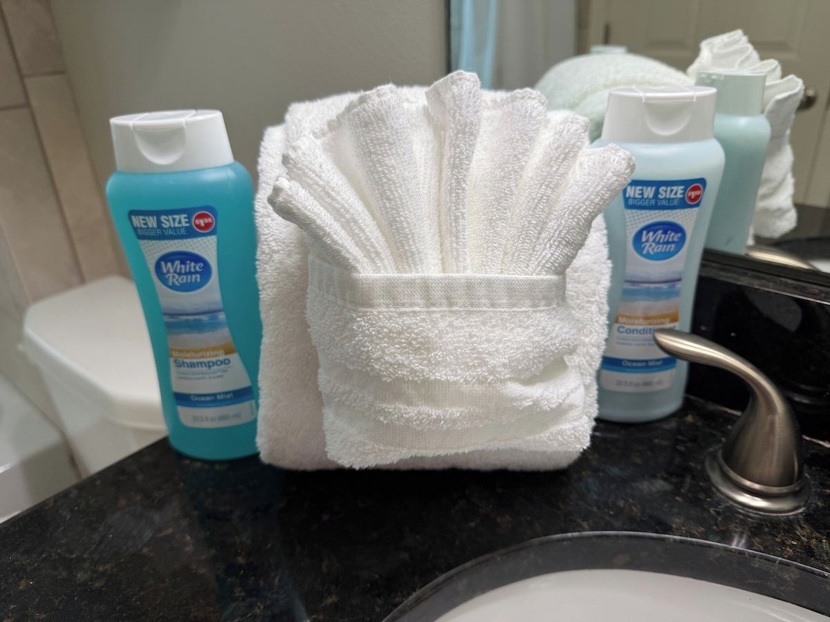 Een badkamer met een witte handdoek gevouwen in een mand, een wastafel en twee flessen White Rain shampoo op het aanrecht.