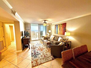 A living room with a sofa, coffee table, and a patterned rug. There is a television on a stand and a lamp on a side table. - Island Time - Gulf Front Retreat (2 Bed 2 Bath) (Pensacola)