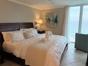 A hotel room with a large bed, a nightstand with a lamp, a painting, and a view of the sea. - Island Time - Gulf Front Retreat (2 Bed 2 Bath) (Pensacola)