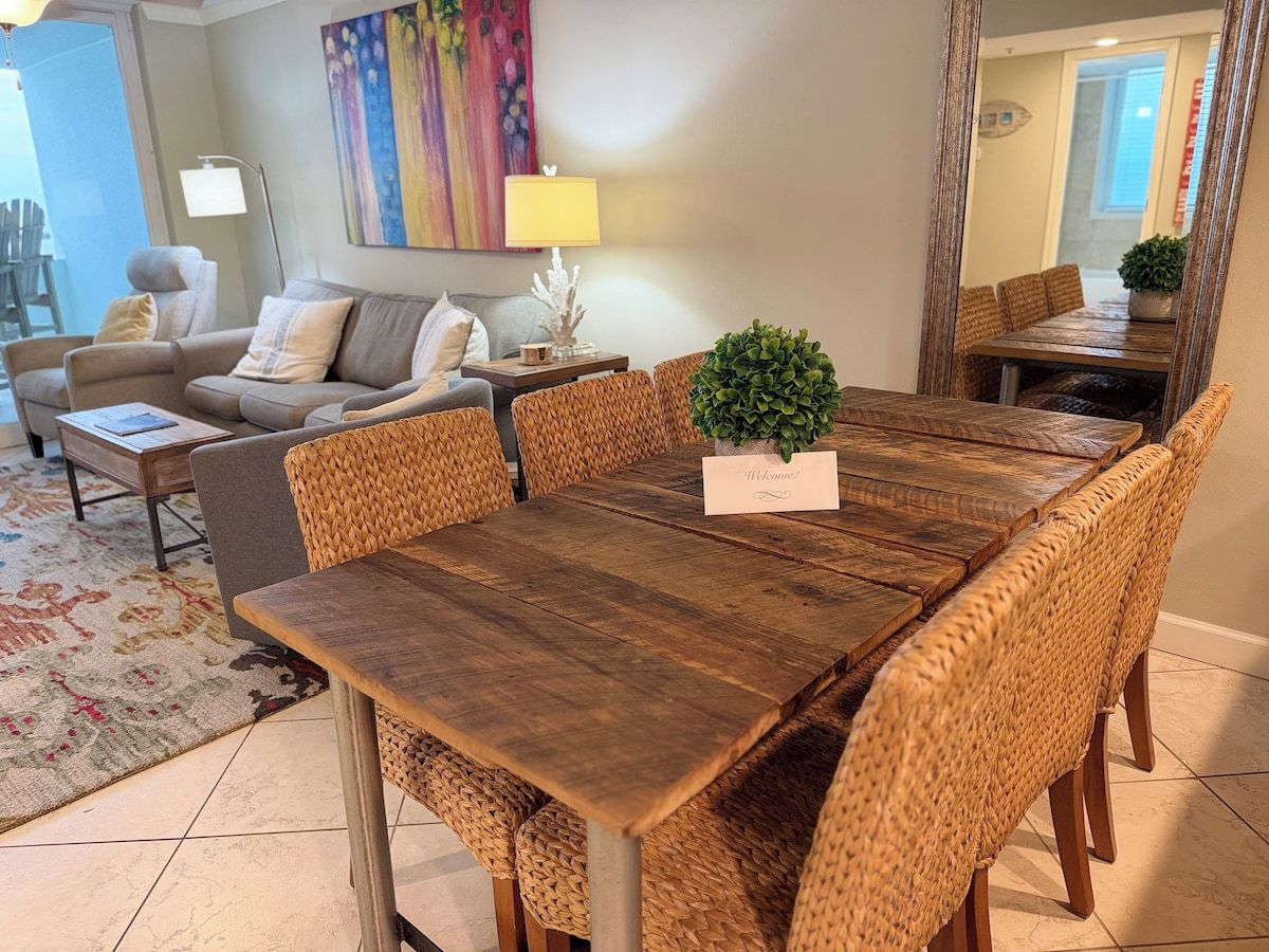 A dining area with a wooden table and wicker chairs, a small potted plant, and a welcome sign.