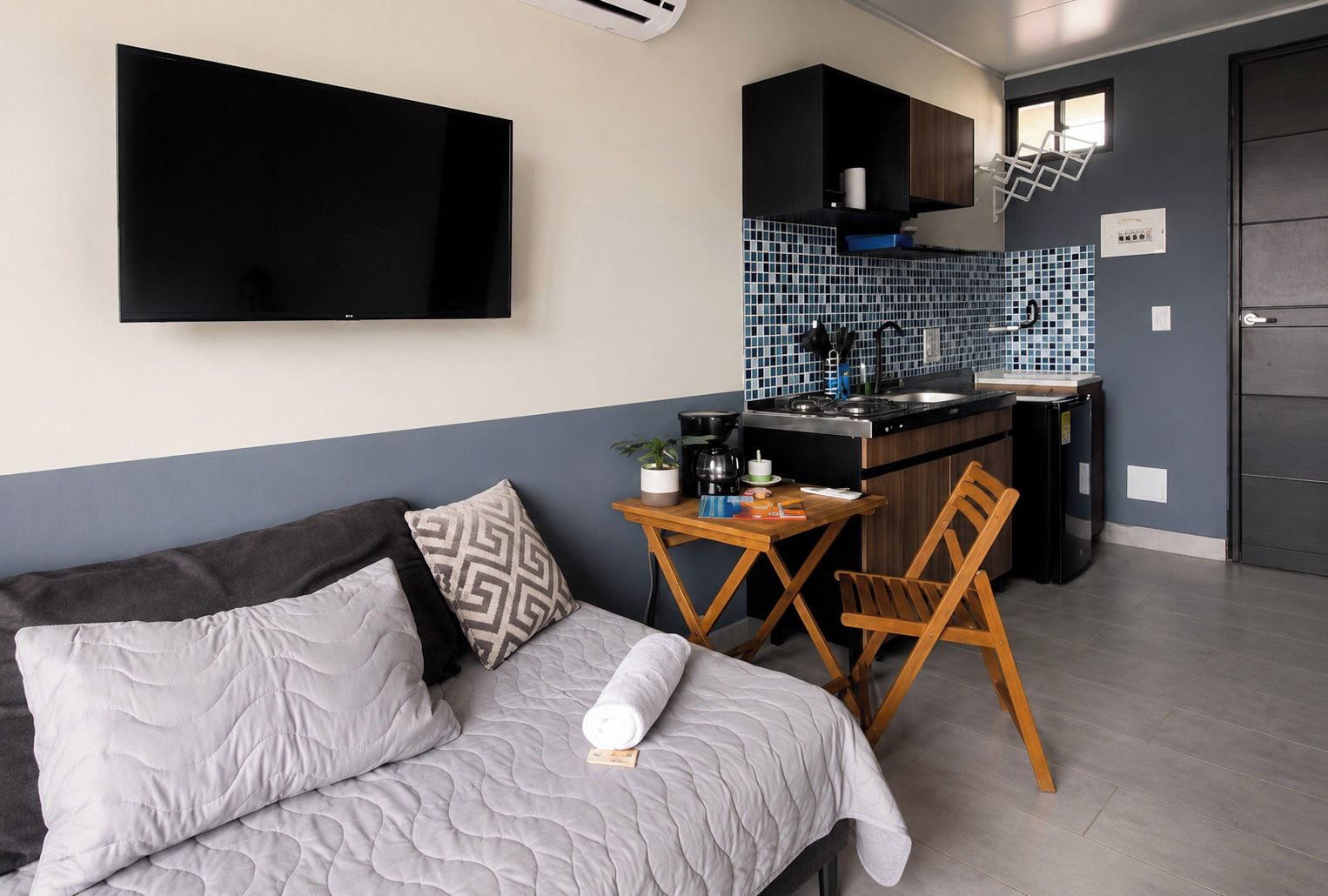 A compact living space with a sofa, a small dining table, a kitchen area with a tiled backsplash, and a flat-screen TV mounted on the wall.