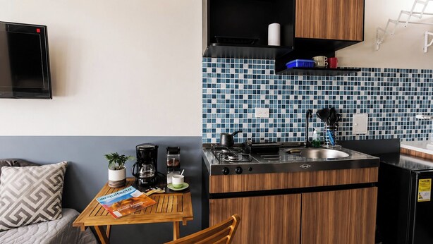 A compact kitchenette with a stove, sink, and a small countertop. There is a dining table with a chair, a coffee maker, and a plant. The room has a blue and white tiled backsplash and wooden cabinets. - Acogedor ApartaSuit amoblado/de 1 dormitorio en Montería con aire acondicionado. (Montería)