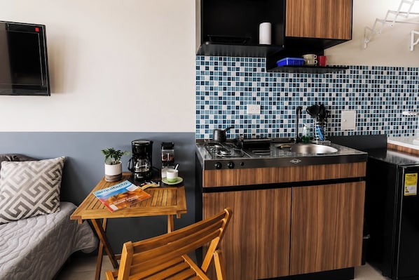 A compact kitchenette with a stove, sink, and a small countertop. There is a dining table with a chair, a coffee maker, and a plant. The room has a blue and white tiled backsplash and wooden cabinets. - Acogedor ApartaSuit amoblado/de 1 dormitorio en Montería con aire acondicionado. (Montería)