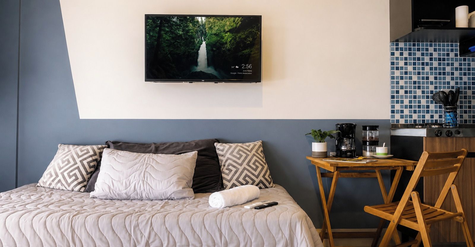 Quarto de hotel com cama, televisão na parede, pequena mesa de madeira e cafeteira, e uma cadeira.