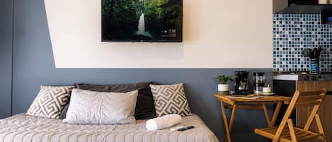 A hotel room with a bed, a TV mounted on the wall, a small wooden table with a coffee maker, and a chair.