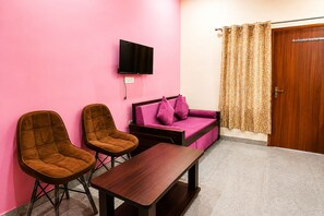 A room with two brown chairs, a wooden table, a pink sofa, a television mounted on the wall, and a door with a patterned curtain. - The Ushmaya Wellness Resort Rishikesh (Rishikesh)