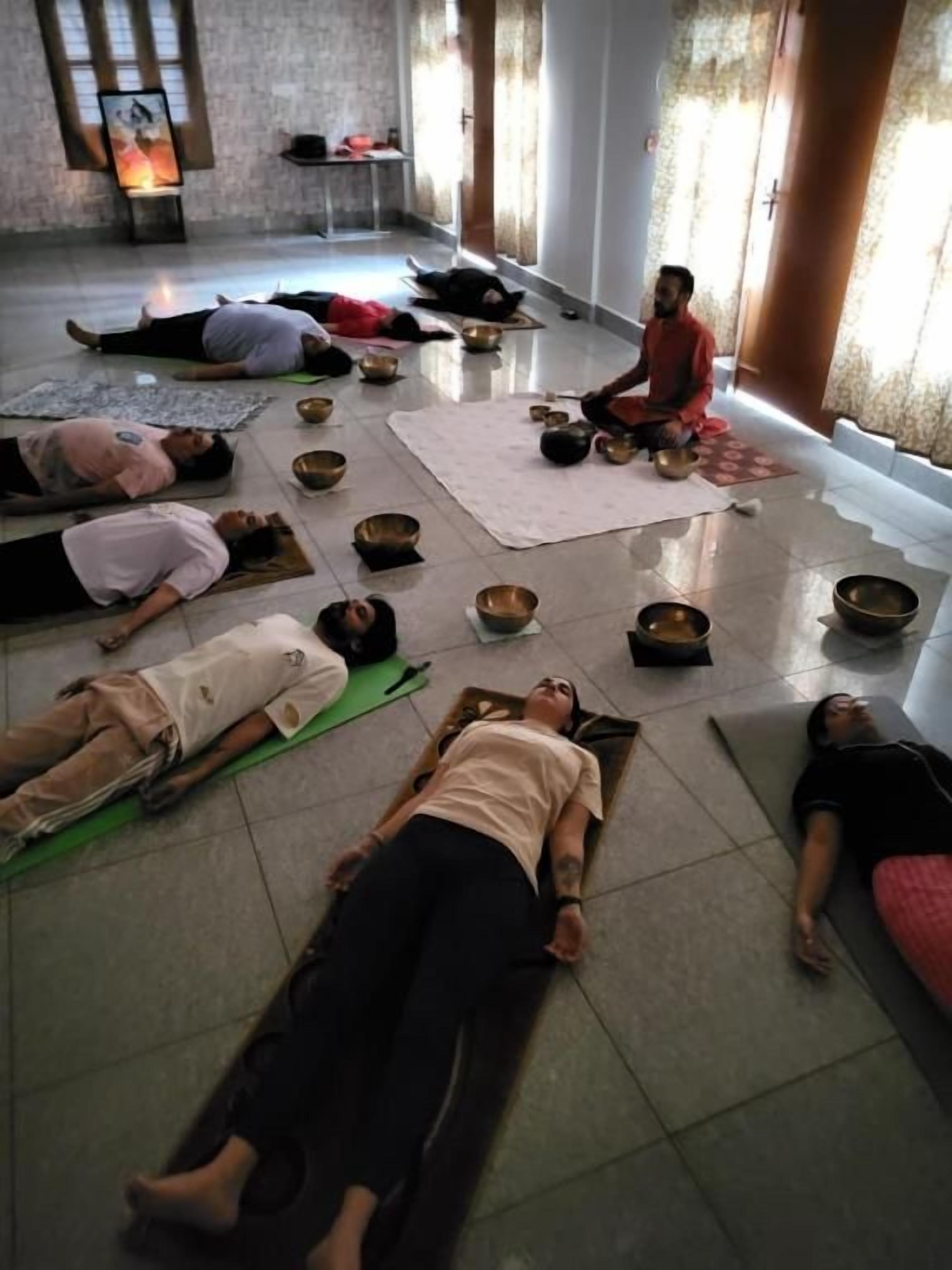 A group of people lying on mats in a room with a tiled floor, some bowls, and a wall with a framed picture.