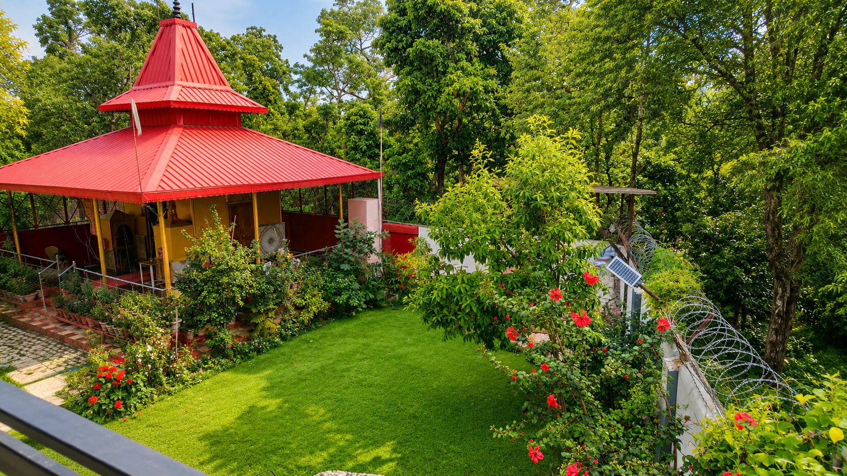 Ein pavillon mit rotem Dach, vor dem ein Garten liegt, umgeben von üppigem Grün und blauem Himmel.