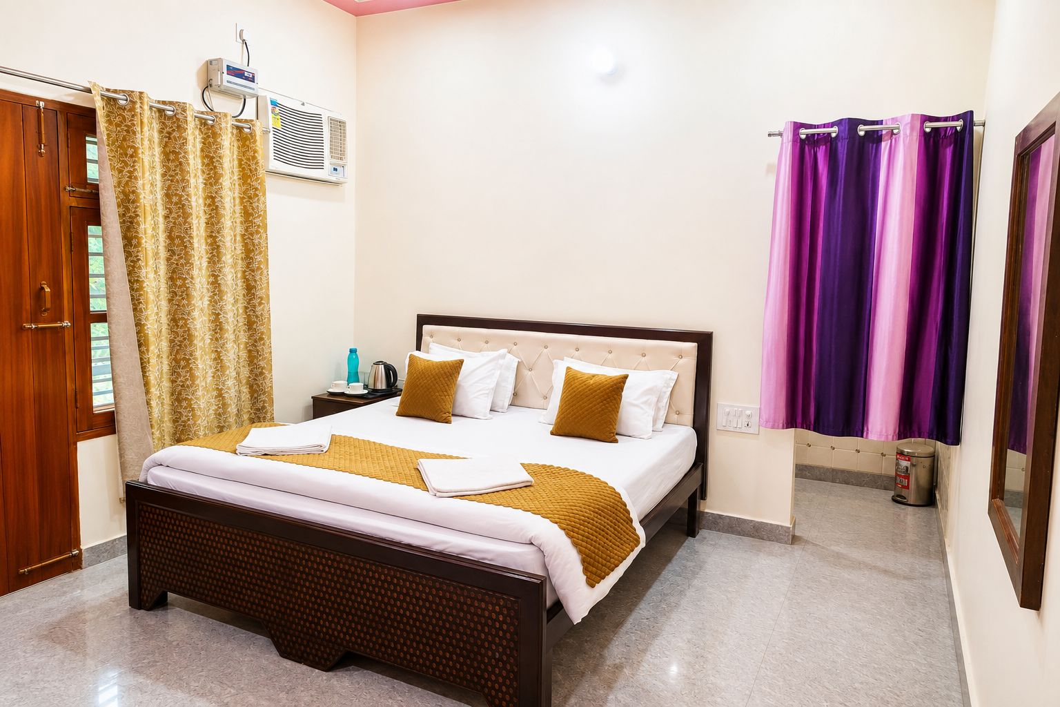 Habitación de hotel con cama, cortinas, unidad de aire acondicionado, mesita de noche con hervidor y un cesto de basura.