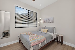 Un dormitorio con cama, mesita de noche, ventana con persianas, lámpara de pared y un cuadro.