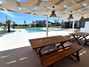 Una terrazza esterna con tavoli e panche in legno, una piscina e una zona coperta con lampade a sospensione.