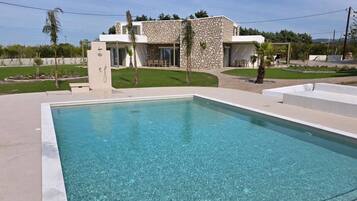 A rectangular swimming pool with clear water, surrounded by a concrete deck. In the background, there is a modern two-story house with stone facade and large windows.