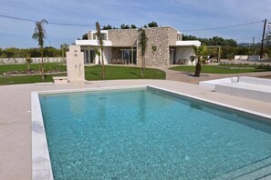 Une piscine rectangulaire à l’eau claire, entourée d’une terrasse en béton. Au fond, on aperçoit une maison moderne à deux étages, dotée d’une façade en pierre et de grandes fenêtres.