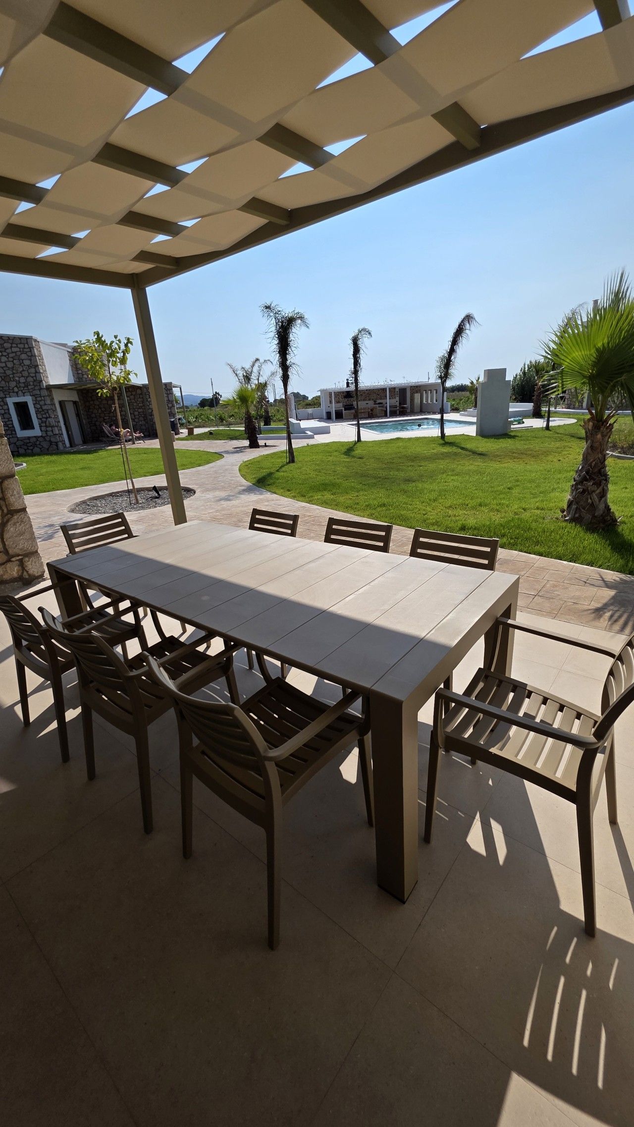 Une terrasse extérieure avec une table rectangulaire et des chaises, entourée d’une structure en forme de pergola.