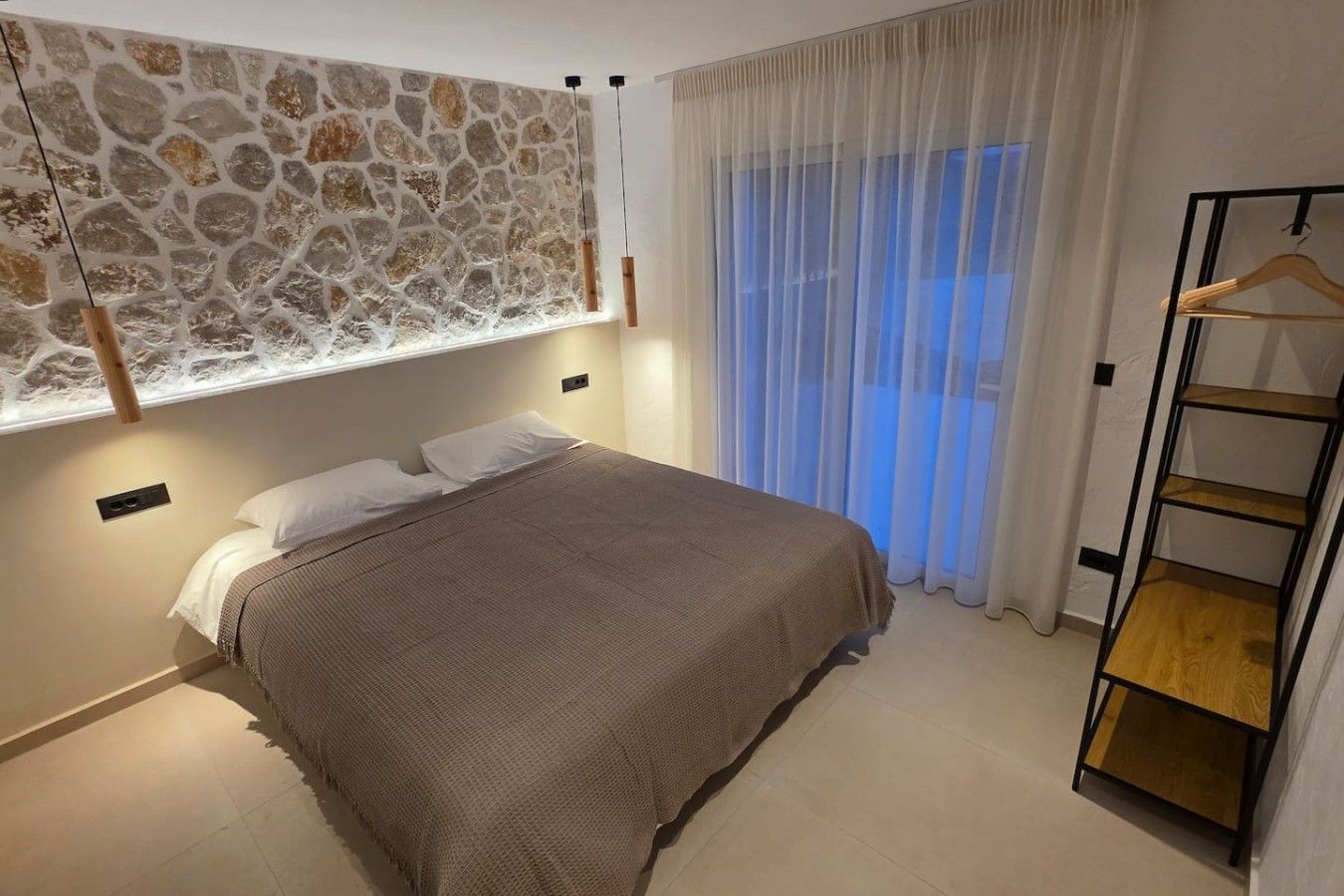 Une chambre avec un lit, un luminaire suspendu, une fenêtre avec des rideaux légers et une étagère en bois.