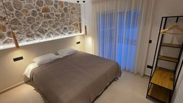 Une chambre avec un lit, un luminaire suspendu, une fenêtre avec des rideaux légers et une étagère en bois.