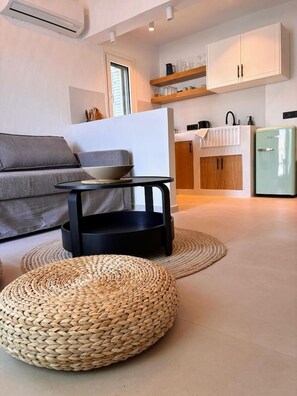 Ein modernes Wohnzimmer mit einer Couch, einem kleinen schwarzen Tisch mit einer Schale, einem gewebten Hocker und einer kleinen Küche im Hintergrund.