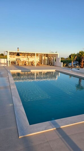 Ein Schwimmbecken mit einer Steinbordsteinkante, eine Terrasse mit Gartenmöbeln und ein Gebäude mit heller Fassade im Hintergrund.