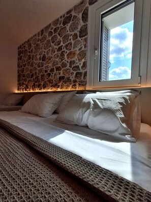 A bed with white bedding, a stone wall, and a window with blinds. - Fanes Breeze Lodges - Lodge Jasmin (Fanes)