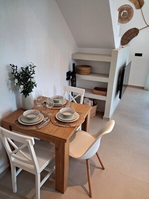 Een eetgedeelte met een houten tafel gedekt voor vier personen, witte stoelen, een vaas met bloemen en een aan de muur bevestigde televisie.