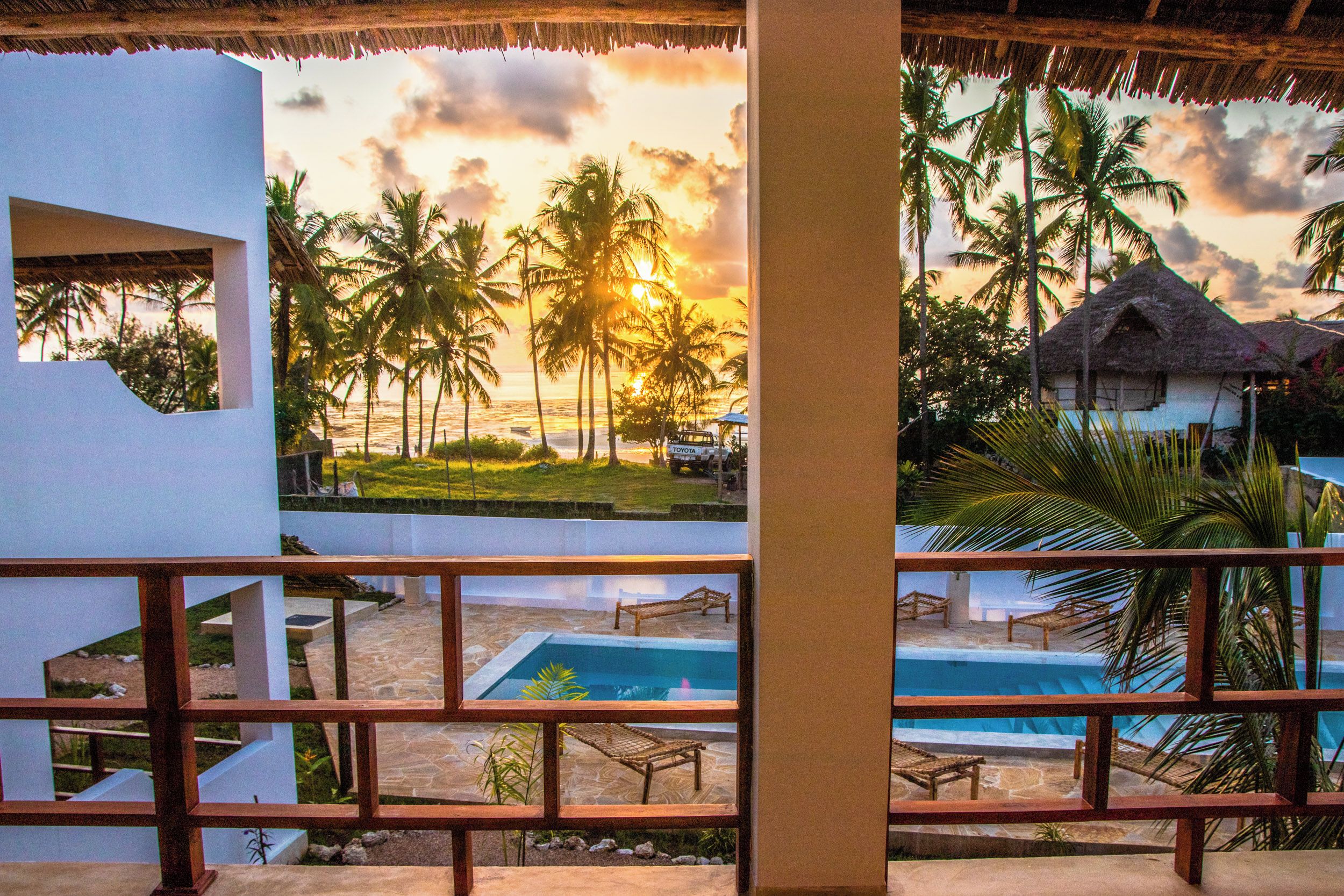 Un resort con piscina, sillas de descanso y vistas a palmeras y al océano al atardecer.