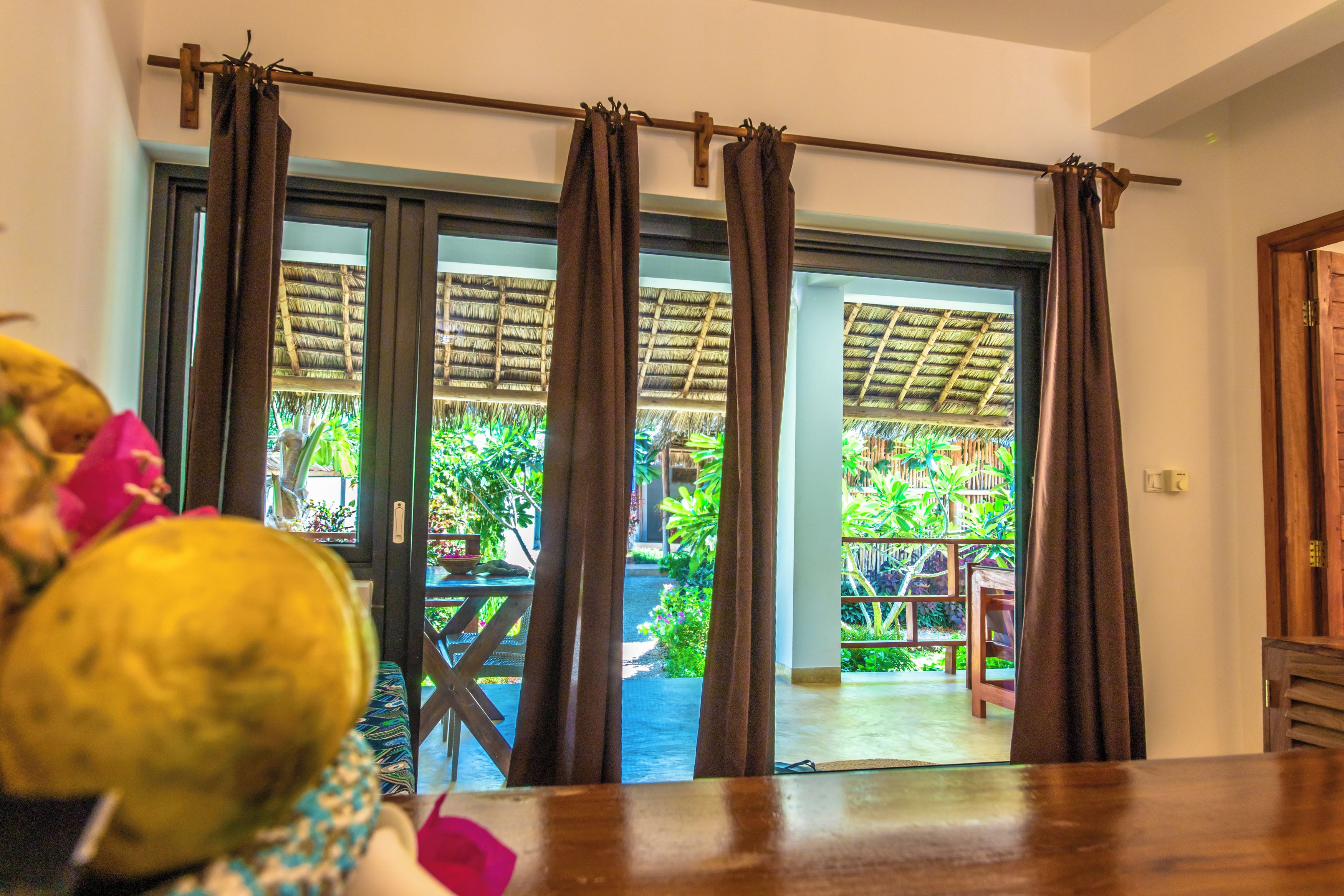 A room with a view of a thatched-roof outdoor area through large windows with curtains.