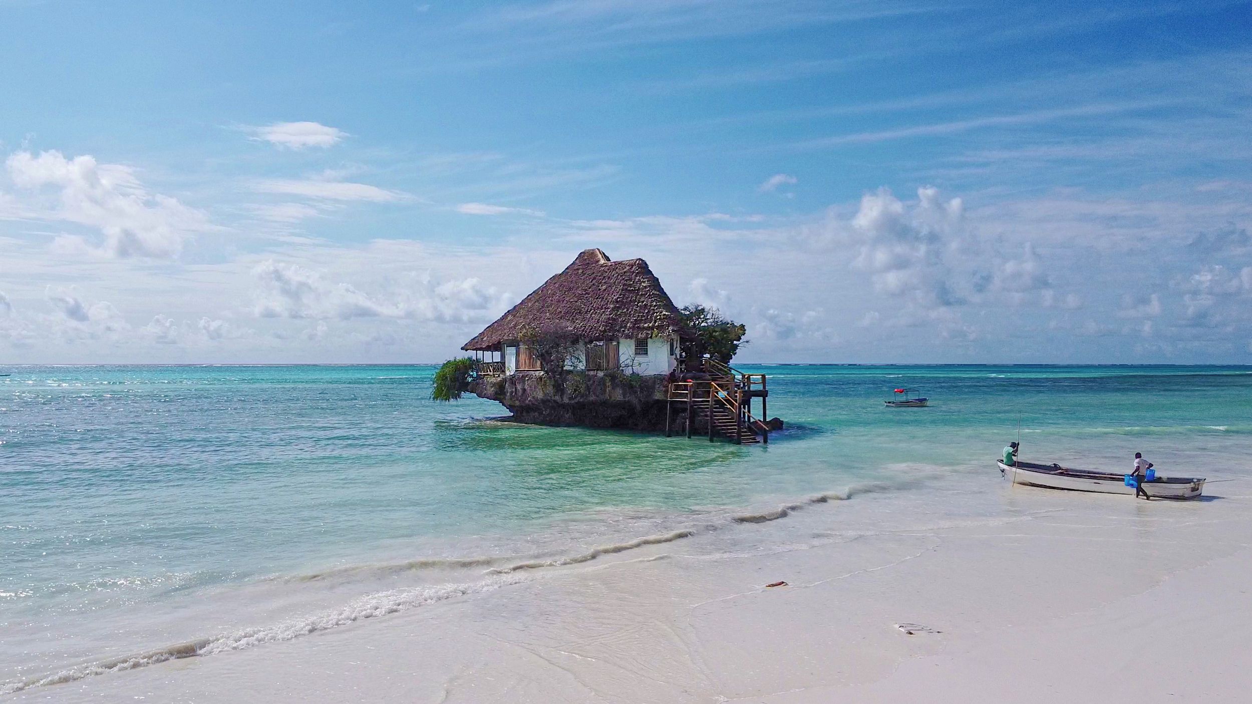 A thatched-roof house on stilts over clear turquoise water, connected to a sandy beach.