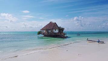 A thatched-roof house on stilts over clear turquoise water, connected to a sandy beach.