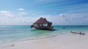 A thatched-roof house on stilts over clear turquoise water, connected to a sandy beach. - Pingwe Beach Apartments (Michamvi)