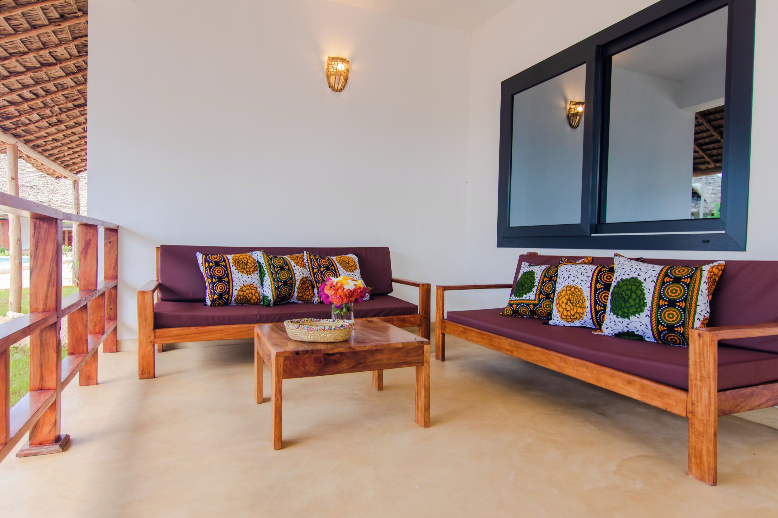 A room with two wooden sofas with patterned cushions, a wooden coffee table with a woven basket and flowers, and a large window with a dark frame.