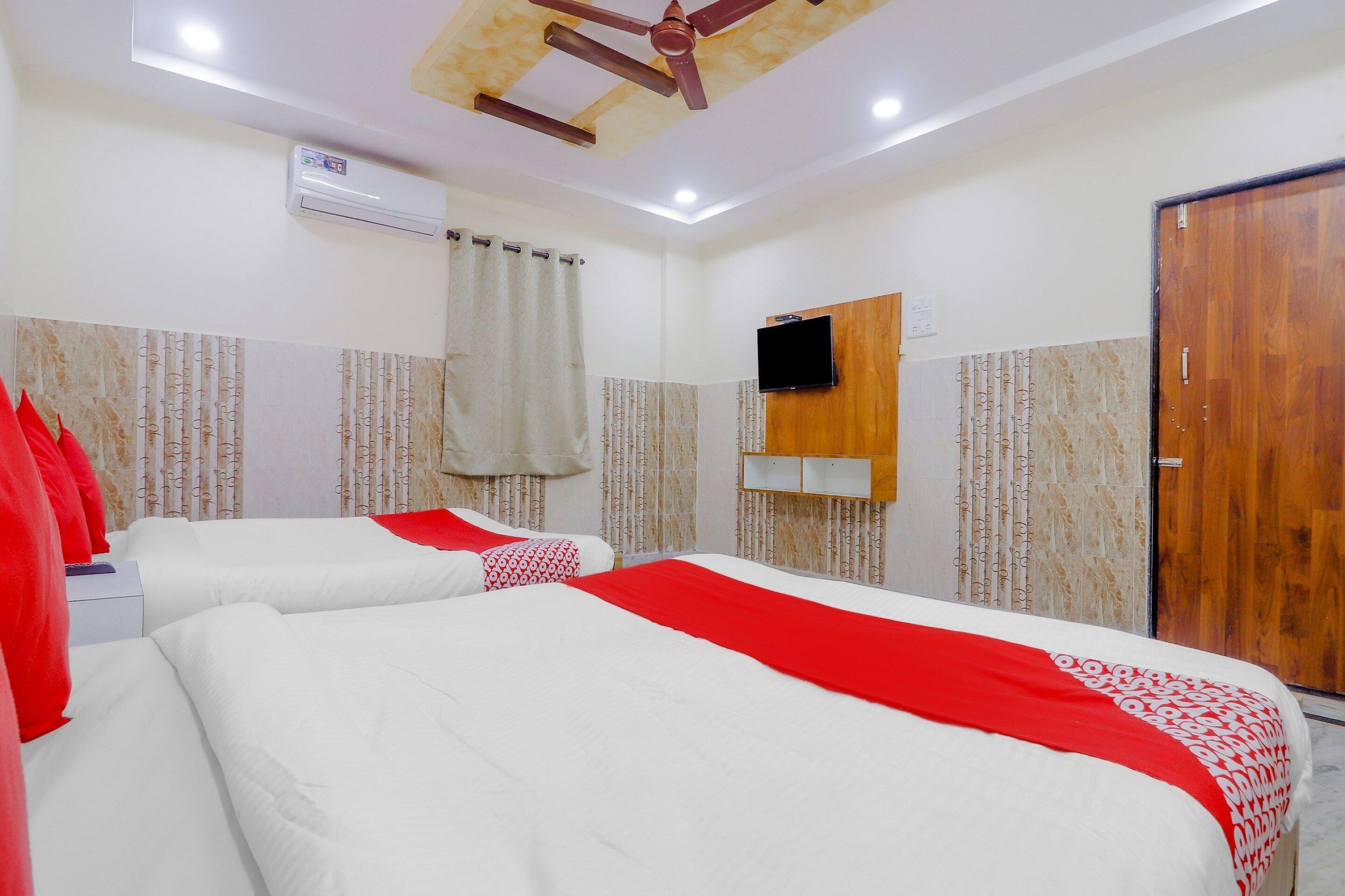 Habitación de hotel con dos camas, ventilador de techo, unidad de aire acondicionado, televisor montado en la pared y puerta de madera.