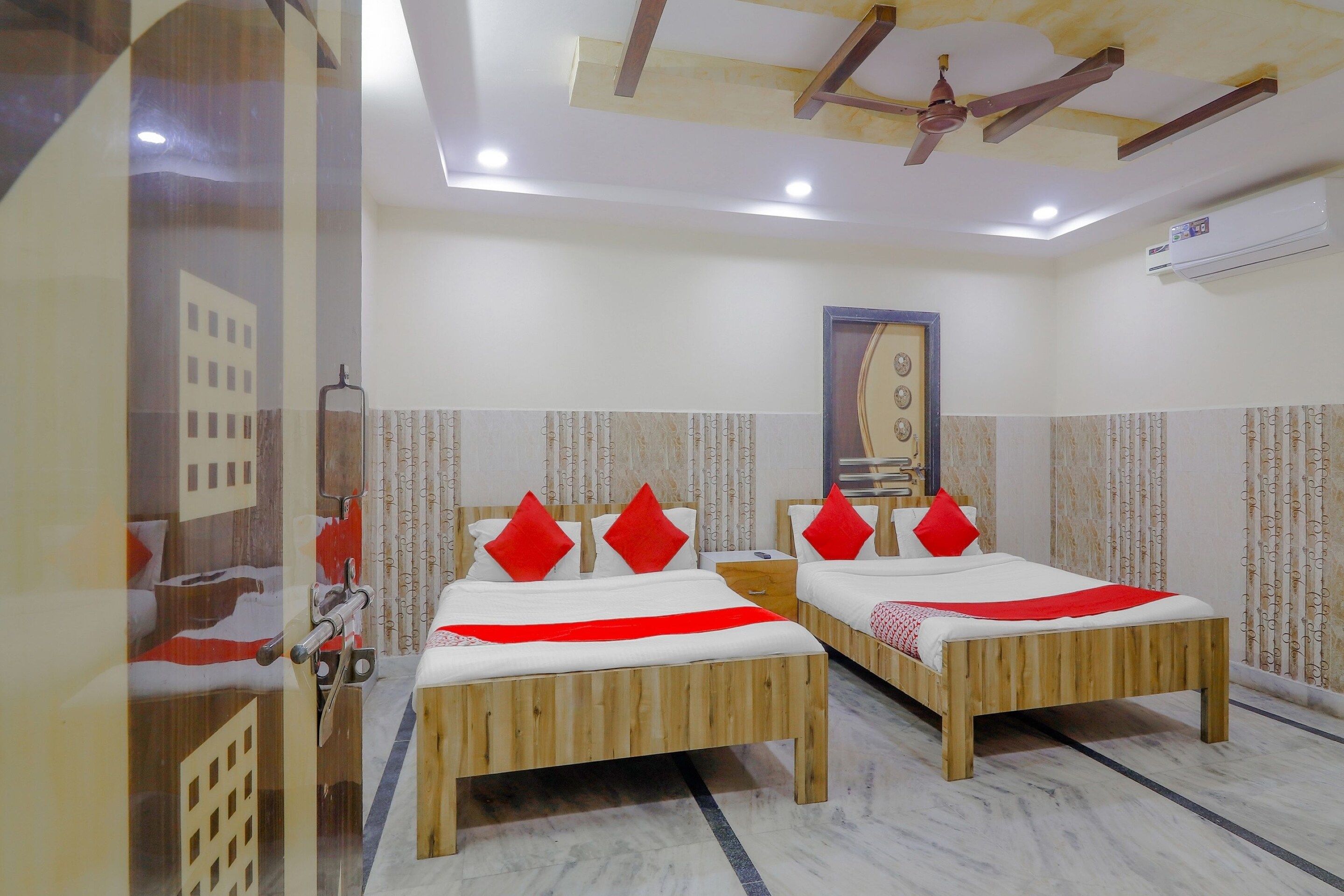 A hotel room with two wooden beds, red pillows, a ceiling fan, and an air conditioning unit.