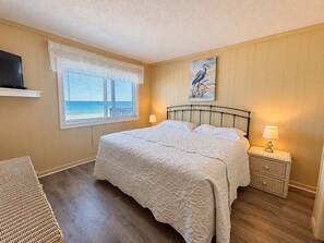 A bedroom with a large bed, a window with blinds, a painting of a bird, and a nightstand with a lamp. - 2 Bedroom Oceanfront Condo, Elevator & Shared Pool in Gareden City! Sea Oaks 206 (Garden City)