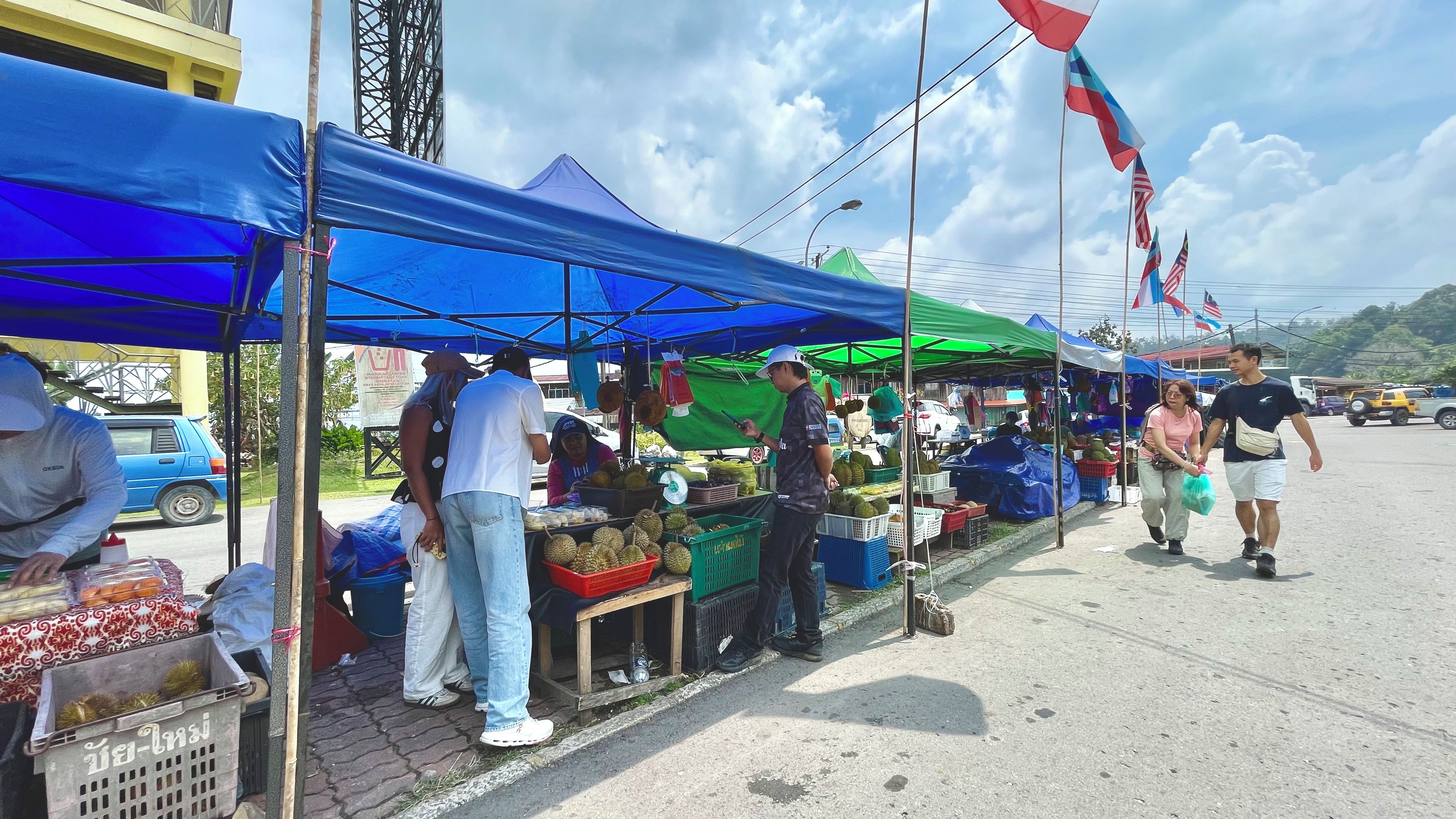 A market stall with durians and other fruits, under blue and green canopies, with flags in the background.