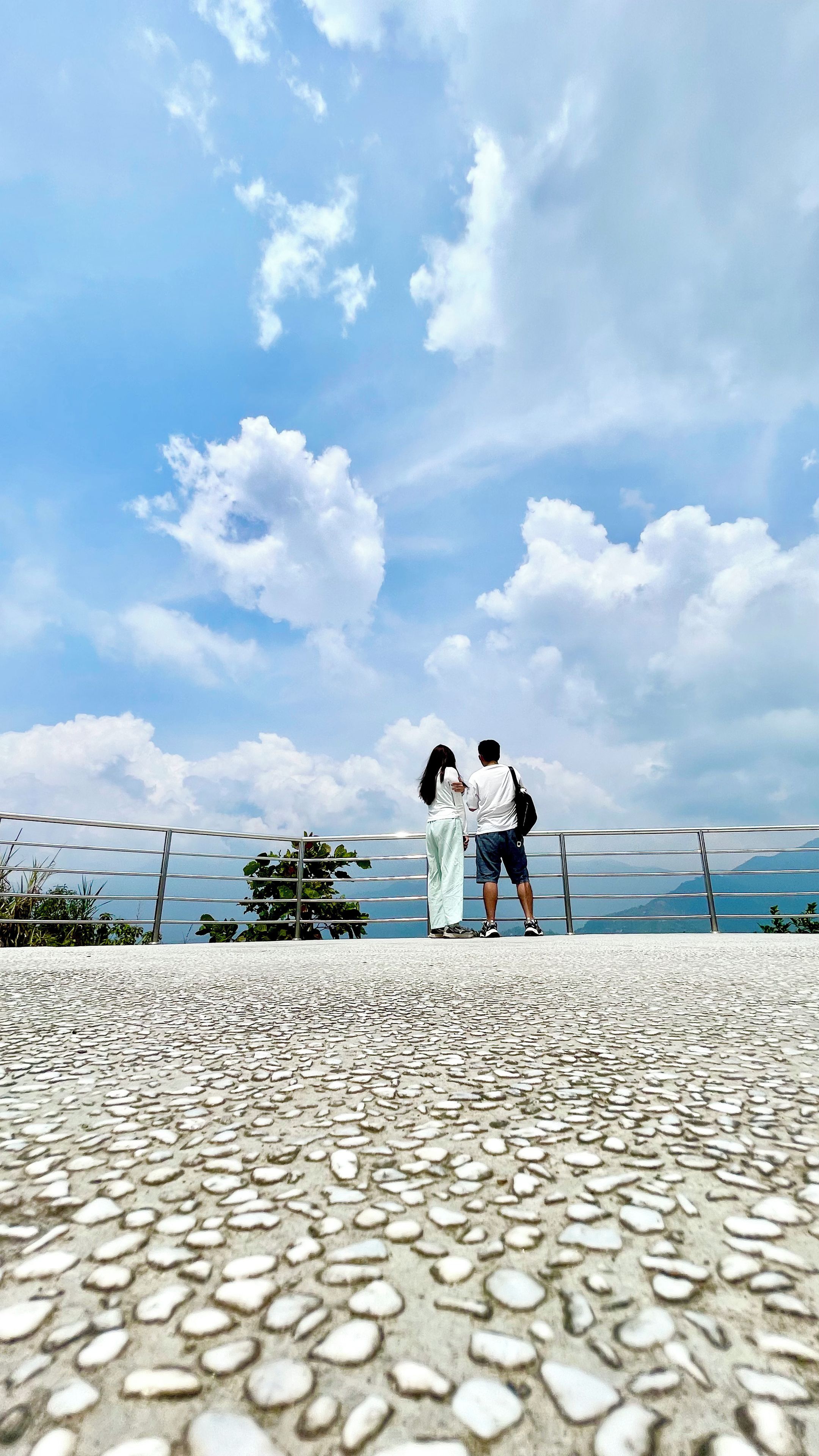 Una pareja está de pie en una zona pavimentada con baranda de metal, contemplando una masa de agua y montañas distantes bajo un cielo parcialmente nublado.